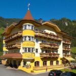 Ein vierstöckiges, gelbes Hotel mit Balkonen vor einer Kulisse aus grünen Hügeln und Bäumen, mit mehreren geparkten Autos davor.