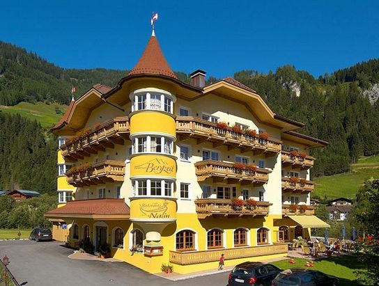 Ein vierstöckiges, gelbes Hotel mit Balkonen vor einer Kulisse aus grünen Hügeln und Bäumen, mit mehreren geparkten Autos davor.