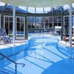 Hallenbadbereich mit Liegestühlen, großen Fenstern und einer Glasdecke, die den Raum mit natürlichem Licht erfüllt.