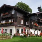 Ein großes Haus im Stil eines dunklen Holzchalets mit Balkonen, die mit Blumenkästen geschmückt sind, und einem Vorgarten in einer grünen, bergigen Gegend.
