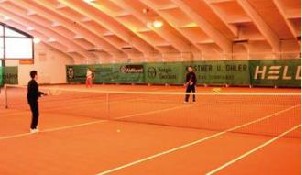 Drei Personen spielen Tennis auf einem Indoor-Sandplatz unter einer weißen Gewölbedecke mit grünen Werbeanzeigen an der Rückwand.