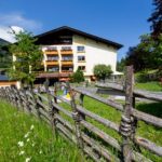 Ein rustikaler Holzzaun führt durch einen grasbewachsenen Hof vor einem mehrstöckigen Gebäude mit Balkonen, umgeben von Bäumen unter einem klaren blauen Himmel.