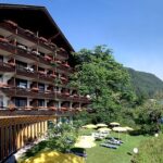 Das vierstöckige Hotel mit Holzbalkonen und Blumenkästen überblickt einen grünen Rasen mit gelben Sonnenschirmen und Liegestühlen vor der Kulisse bewaldeter Hügel.