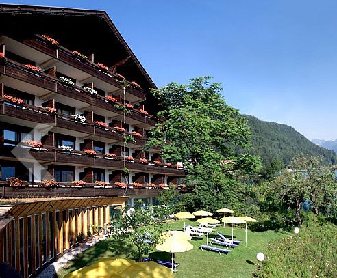 Das vierstöckige Hotel mit Holzbalkonen und Blumenkästen überblickt einen grünen Rasen mit gelben Sonnenschirmen und Liegestühlen vor der Kulisse bewaldeter Hügel.