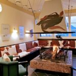 Eine gemütliche Lounge mit mit Kuhfell gepolsterten Bänken, einem zentralen offenen Kamin, abstrakter Kunst und großen Fenstern mit Blick auf die Berge.