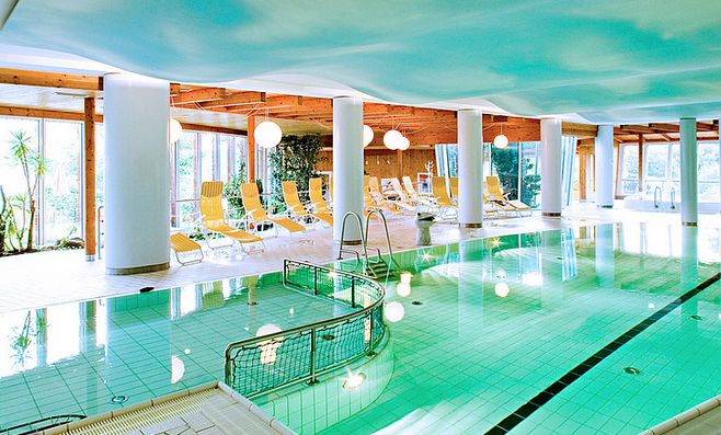 Hallenbad mit klarem Wasser, Liegestühlen und großen Fenstern, die natürliches Licht hereinlassen; weiße Säulen und eine Holzdecke sind sichtbar.