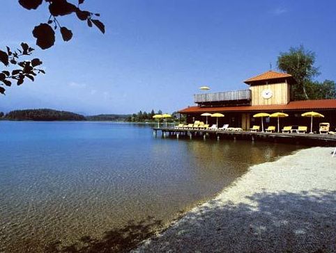 Ein Gebäude am See mit einer Terrasse, gelben Sonnenschirmen und Liegestühlen liegt an einem klaren, ruhigen Wasser und einem Kiesstrand unter blauem Himmel.