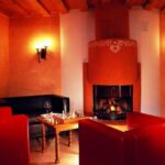 Ein gemütliches Zimmer mit roten Sesseln, einem schwarzen Sofa, einem Holztisch mit Gläsern und einem Schachbrett und einem beleuchteten Kamin in einem orangefarbenen Schornstein.
