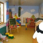 Ein Kinderspielzimmer mit buntem Spielzeug, einer Kreidetafel mit der Aufschrift "HALLO KINDER", einem großen weißen Stoffbären und blauen Vorhängen mit Cartoon-Motiven.