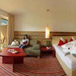 Hotelzimmer mit einem Doppelbett, roten und weißen Kissen, einer kleinen Sitzecke, gestreiftem Teppich, großen Fenstern und einer Frau, die auf dem Sofa in der Nähe des Balkons mit Blick ins Grüne sitzt.