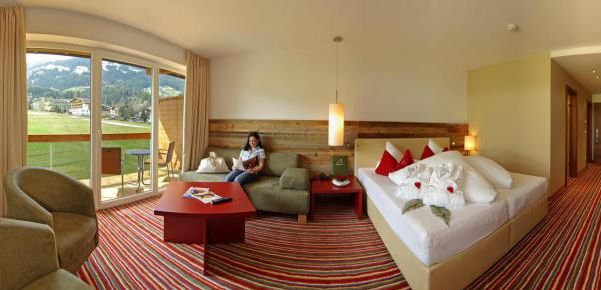 Hotelzimmer mit einem Doppelbett, roten und weißen Kissen, einer kleinen Sitzecke, gestreiftem Teppich, großen Fenstern und einer Frau, die auf dem Sofa in der Nähe des Balkons mit Blick ins Grüne sitzt.