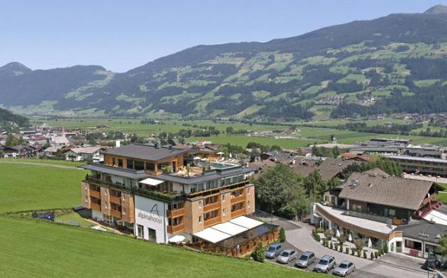 Ein mehrstöckiges Hotelgebäude steht neben einer grünen Wiese mit Bergen und einem Dorf im Hintergrund unter einem klaren blauen Himmel.