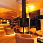 Moderne Hotellounge mit warmer Beleuchtung, zentralem hängendem Kamin, braunen Sesseln und großen Fenstern; im Hintergrund ist ein Weihnachtsbaum zu sehen.