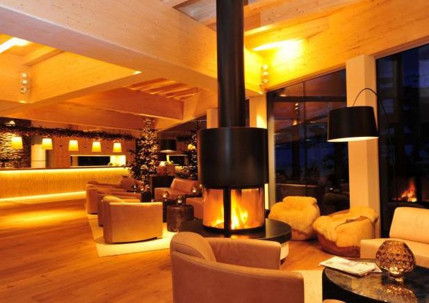 Moderne Hotellounge mit warmer Beleuchtung, zentralem hängendem Kamin, braunen Sesseln und großen Fenstern; im Hintergrund ist ein Weihnachtsbaum zu sehen.