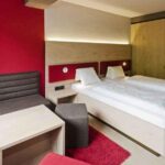 Modernes Hotelzimmer mit einem Doppelbett, zwei weißen Kissen, einem grauen Sofa, einem kleinen Tisch, einer roten Akzentwand und einem Teppich.