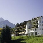 Ein großes, mehrstöckiges Hotel mit Balkonen und einem dunklen Dach liegt auf einem grasbewachsenen Hügel, mit Bergen und Bäumen im Hintergrund unter einem klaren Himmel.