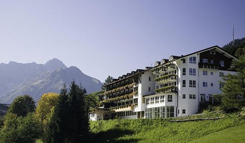 Ein großes, mehrstöckiges Hotel mit Balkonen und einem dunklen Dach liegt auf einem grasbewachsenen Hügel, mit Bergen und Bäumen im Hintergrund unter einem klaren Himmel.