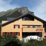 Ein dreistöckiges Gebäude mit der Aufschrift "Hotel Post" und roten Fensterläden vor einer Kulisse aus grünen Bergen und einem teilweise bewölkten Himmel.