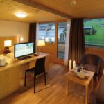 Ein gemütliches Hotelzimmer mit Holzmöbeln, einem Bett, einem Schreibtisch mit Fernseher, einem Sofa, einem Couchtisch mit Wein und großen Fenstern mit Blick auf eine grüne Landschaft.