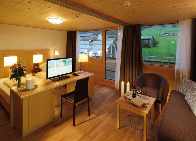 Ein gemütliches Hotelzimmer mit Holzmöbeln, einem Bett, einem Schreibtisch mit Fernseher, einem Sofa, einem Couchtisch mit Wein und großen Fenstern mit Blick auf eine grüne Landschaft.