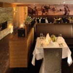 Modernes Restaurantinterieur mit ordentlich gedeckten Tischen, braunen Ledersesseln, einer Akzentwand aus Stein mit Kamin und einem Wandgemälde mit Außenaufnahmen an der Rückwand.