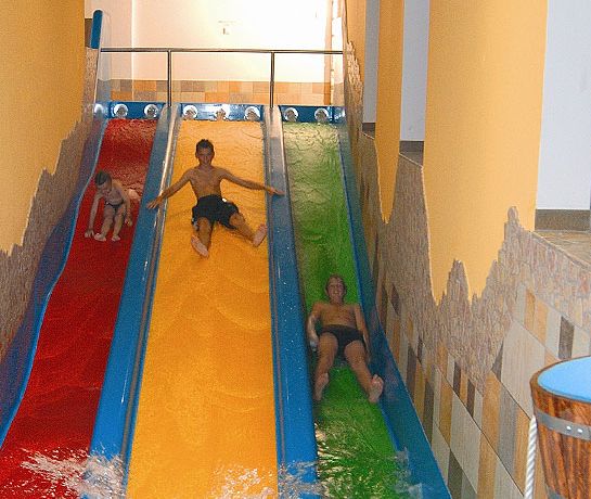 Drei Personen rutschen auf parallelen Wasserrutschen in den Farben Rot, Gelb und Grün hinunter, umgeben von hellbraunen Kachelwänden und Säulen.