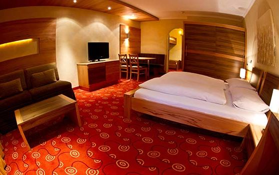 Ein Hotelzimmer mit einem Doppelbett, einem Sofa, einem Fernseher auf einer Holzkommode, einem kleinen Esstisch mit Stühlen und einem gemusterten roten Teppichboden.