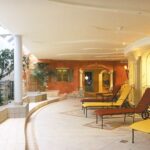 Ein geräumiger Wellnessbereich im Innenbereich mit Liegestühlen, Topfpflanzen, Steinakzenten und architektonischen Bogenelementen bei sanfter Beleuchtung.