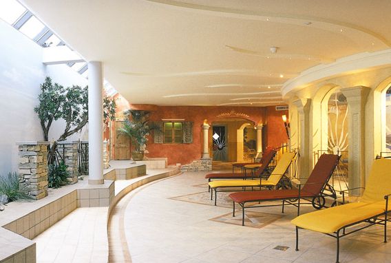 Ein geräumiger Wellnessbereich im Innenbereich mit Liegestühlen, Topfpflanzen, Steinakzenten und architektonischen Bogenelementen bei sanfter Beleuchtung.