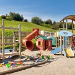 Spielplatz im Freien mit Klettergerüsten, Rutschen, einem Spielhaus und verstreutem bunten Spielzeug auf Sand, eingebettet in eine eingezäunte Rasenfläche mit Hügeln im Hintergrund.