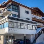 Ein modernes mehrstöckiges Hotel mit Balkonen und hölzernen Akzenten, umgeben von Schnee, mit geparkten Autos davor und einem blauen Himmel darüber.