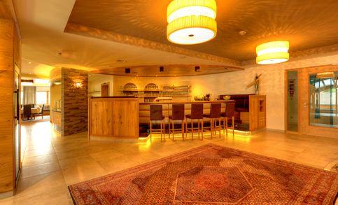 Ein warm beleuchteter Barbereich mit Holzmöbeln, hohen Stühlen, Regalen mit Flaschen und einem großen gemusterten Teppich auf einem gefliesten Boden.