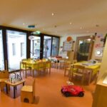 Ein hell erleuchteter Krippenraum mit kleinen Tischen und Stühlen, Kinderspielzeug und Malutensilien, die ordentlich im Raum verteilt sind.