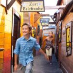 Zwei Kinder rennen lächelnd durch eine bunte Gasse mit Holzhäusern, vorbei an Schildern mit der Aufschrift "Zur Brieftaube" und "Polizei".
