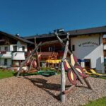 Ein weißes Gästehaus mit Holzbalkonen und Blumenkästen steht hinter einem Spielplatz mit Schaukel auf einer grasbewachsenen Wiese, und im Hintergrund sind die Berge zu sehen.