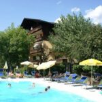 An einem sonnigen Tag faulenzen Menschen auf Stühlen und schwimmen in einem von Bäumen und einem mehrstöckigen Gebäude umgebenen Freibad.