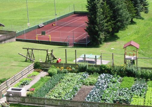 Luftaufnahme eines eingezäunten Gemüsegartens, eines Kinderspielplatzes mit Schaukeln und einer Rutsche und eines eingezäunten Tennisplatzes im Hintergrund.