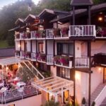 Dreistöckiges Gebäude mit Balkonen, die mit Blumenkästen geschmückt sind; darunter ein Essbereich im Freien mit Tischen unter einer Pergola; umgeben von Grünanlagen.