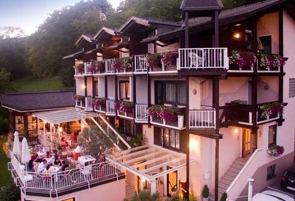 Dreistöckiges Gebäude mit Balkonen, die mit Blumenkästen geschmückt sind; darunter ein Essbereich im Freien mit Tischen unter einer Pergola; umgeben von Grünanlagen.