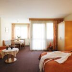 Hotelzimmer mit Doppelbett, Sofa, Kleiderschrank, kleinem Esstisch mit Stühlen und großen Fenstern mit Vorhängen; in warmen Tönen gehaltenes Dekor und ordentliches Mobiliar.