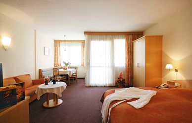 Hotelzimmer mit Doppelbett, Sofa, Kleiderschrank, kleinem Esstisch mit Stühlen und großen Fenstern mit Vorhängen; in warmen Tönen gehaltenes Dekor und ordentliches Mobiliar.