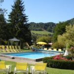 Swimmingpool im Freien mit gelben Liegestühlen, grünem Rasen, Bäumen und Hügeln im Hintergrund unter einem klaren blauen Himmel.