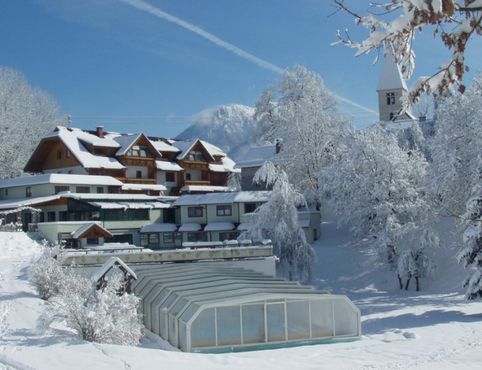 Schneebedeckte Gebäude und Bäume in einer winterlichen Dorfumgebung, mit einem Berg im Hintergrund und einem überdachten Gebäude im Vordergrund.