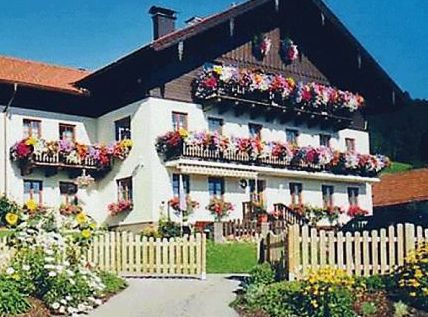 Ein großes Haus mit hölzernen Balkonen, die mit bunten Blumen geschmückt sind; ein Garten und ein Holzzaun davor unter einem klaren blauen Himmel.