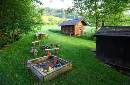 Ein grasbewachsener Hof mit einem mit Spielzeug gefüllten Sandkasten, Holzbänken um eine Feuerstelle und zwei kleinen Holzhütten, umgeben von Bäumen und Feldern.