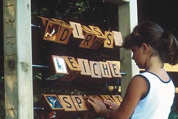 Ein Mädchen dreht auf einer Außenkonstruktion Holzklötze mit Buchstaben und bildet so deutsche Wörter in drei Reihen.