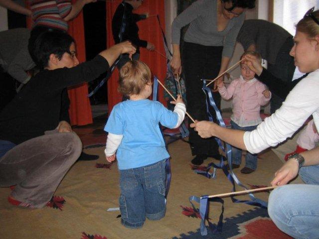 Mehrere Erwachsene und kleine Kinder sind in einem Raum versammelt, halten Stöcke mit blauen Bändern in der Hand und üben eine Gruppenaktivität auf einem Teppichboden aus.