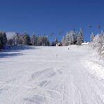Eine schneebedeckte Skipiste mit Skispuren, umgeben von Bäumen und mehreren Skiliften im Hintergrund unter einem klaren blauen Himmel.