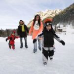 Eine vierköpfige Familie beim Schlittschuhlaufen im Freien an einem verschneiten Tag mit Bergen und Bäumen im Hintergrund.