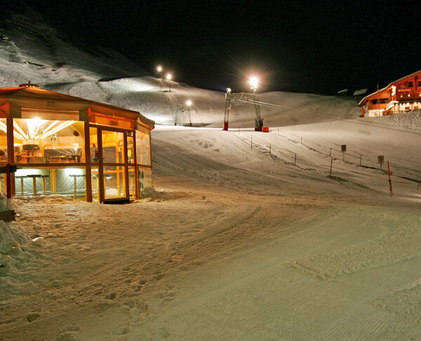 Schneebedecktes Skigebiet bei Nacht mit einem beleuchteten Glasrestaurant und einer Holzhütte, beleuchteten Pisten und sichtbaren Skiliftanlagen.